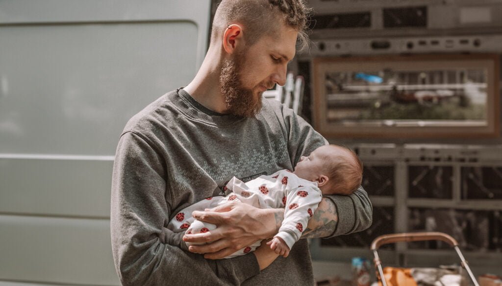 Wojtek z miłością wpatruje się w kilkumiesięczną Zoję. W tle nasz kamper w ogródku mamy, który jest w trakcie budowy. To moment pełen uczucia i oczekiwania na wspólne podróże.